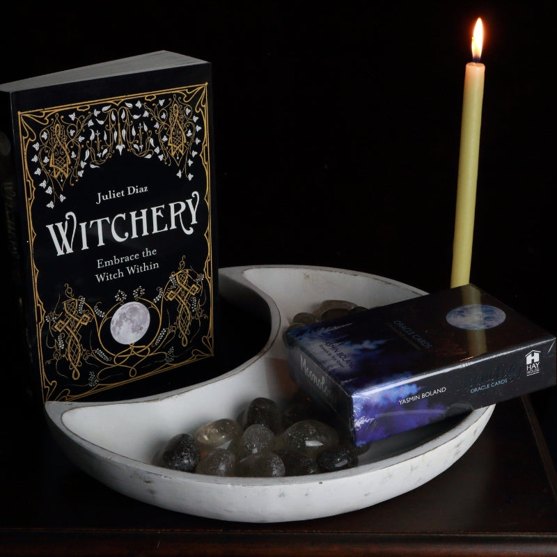 whitewash crescent moon shaped bowl filled with smoky quartz crystals and holding a moonology oracle deck, in front of a juliet Diaz book called "Witchery" and a beeswax prayer candle