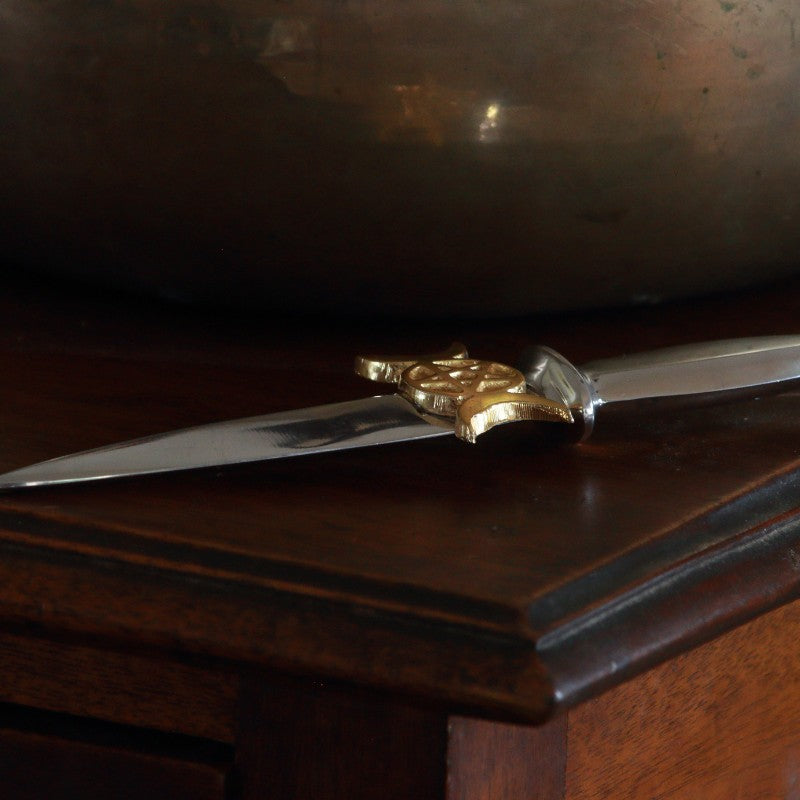 aluminium athame/ dagger with a gold pentacle and two gold crescent moons at each side beneath the hilt laying on a wooden table
