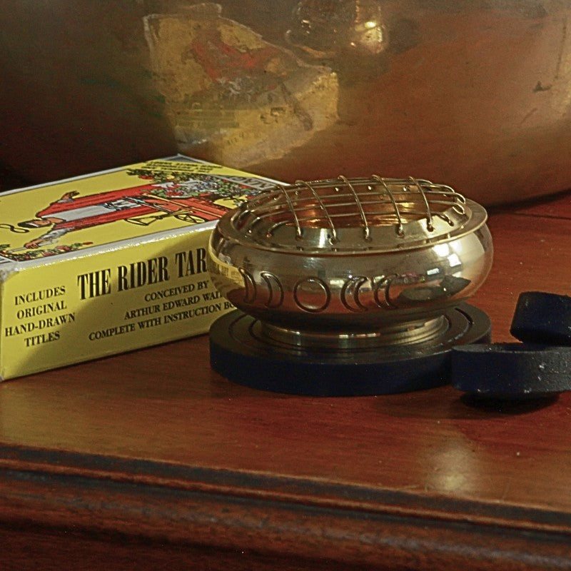 Brass charcoal incense burner engraved with the phases of the moon, sitting on a wooden base plate next to charcoal discs and a rider waite smith tarot deck