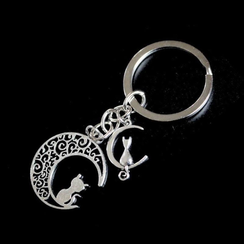 silver coloured key ring with a a large and small crescent moon each with a cat sitting on the moon, joined to a silver chain which links onto a silver ring on a black background