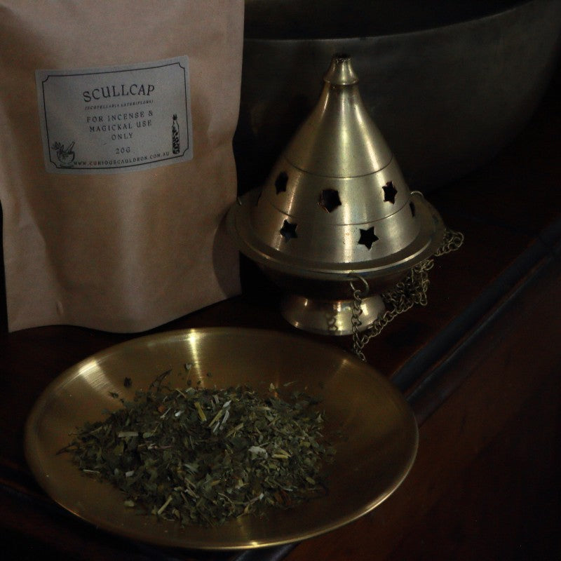 Brass bowl with dried herbs, brass cone incense burner, and Scullcap packaging on a wooden surface.