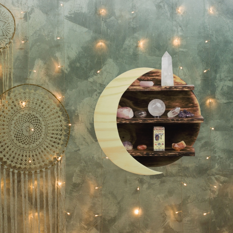 moon shaped crystal shelf containig crystals and meditation essential oil, hung on a green wall with fairy lights and a dream catcher