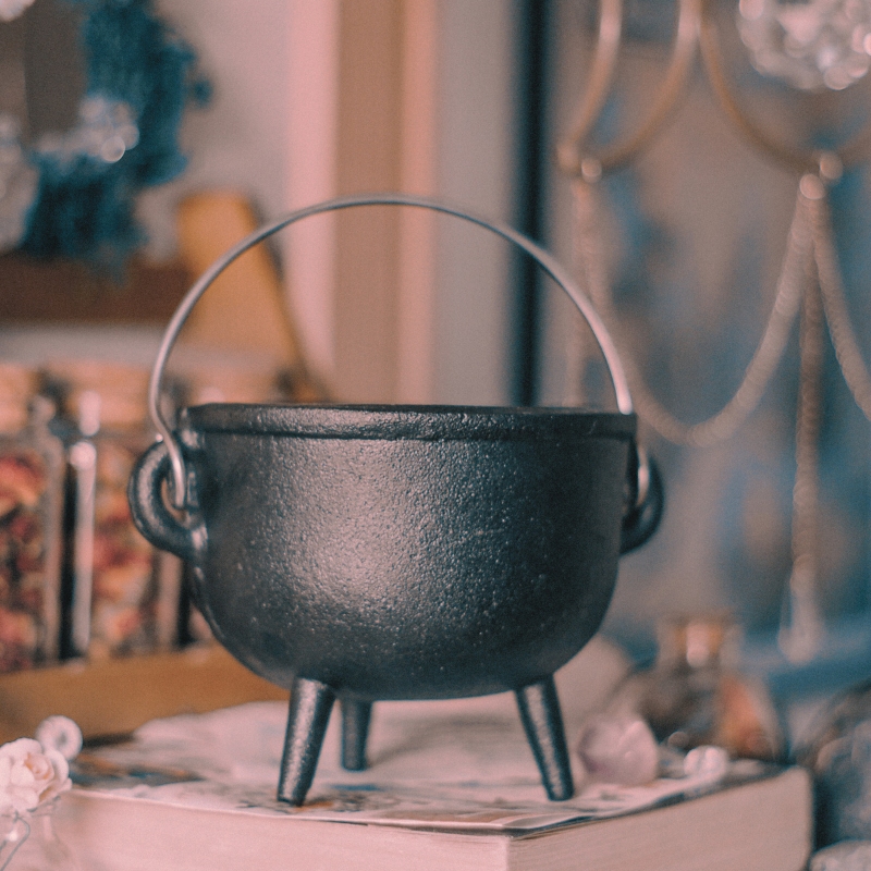 A black cauldron with a silver handle sitting on top of a thick book with glass jars of herbs in the background