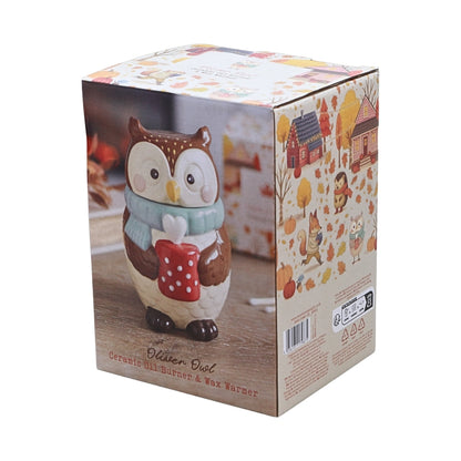 An owl-shaped ceramic oil burner and wax warmer shown in its retail packaging box. The box features a printed illustration of the owl holding a red mug, with autumn-themed graphics on the side panel.