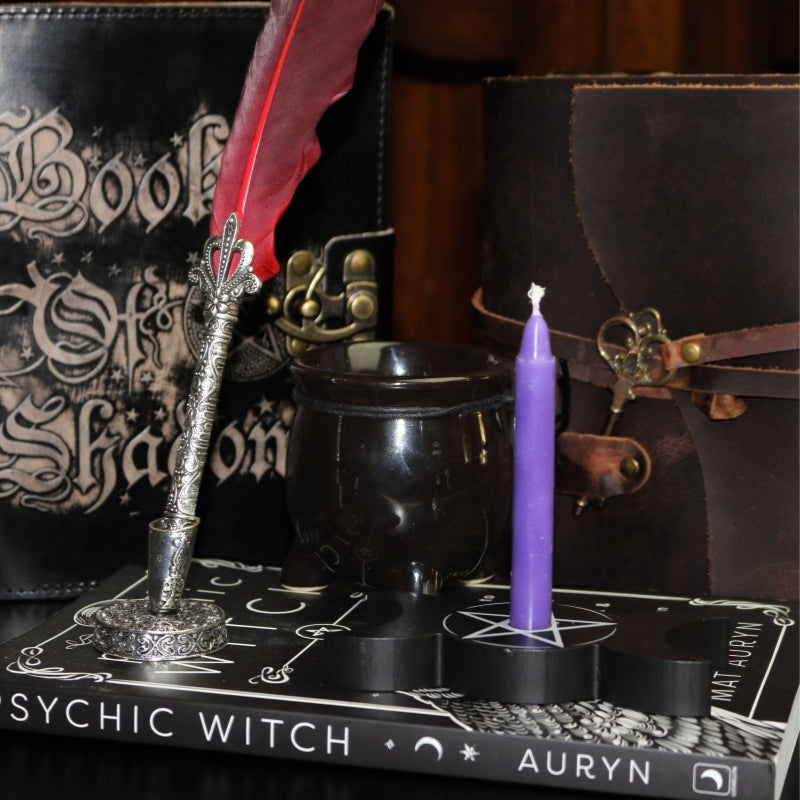 Red feather dip pen with silver handle, sitting in a silver pen holder on top of a book "psychic witch", next to a black ceramic cauldron candle, triple moon spell candle holder with a purple spell candle, in front of a key closure leather journal and book of shadows 