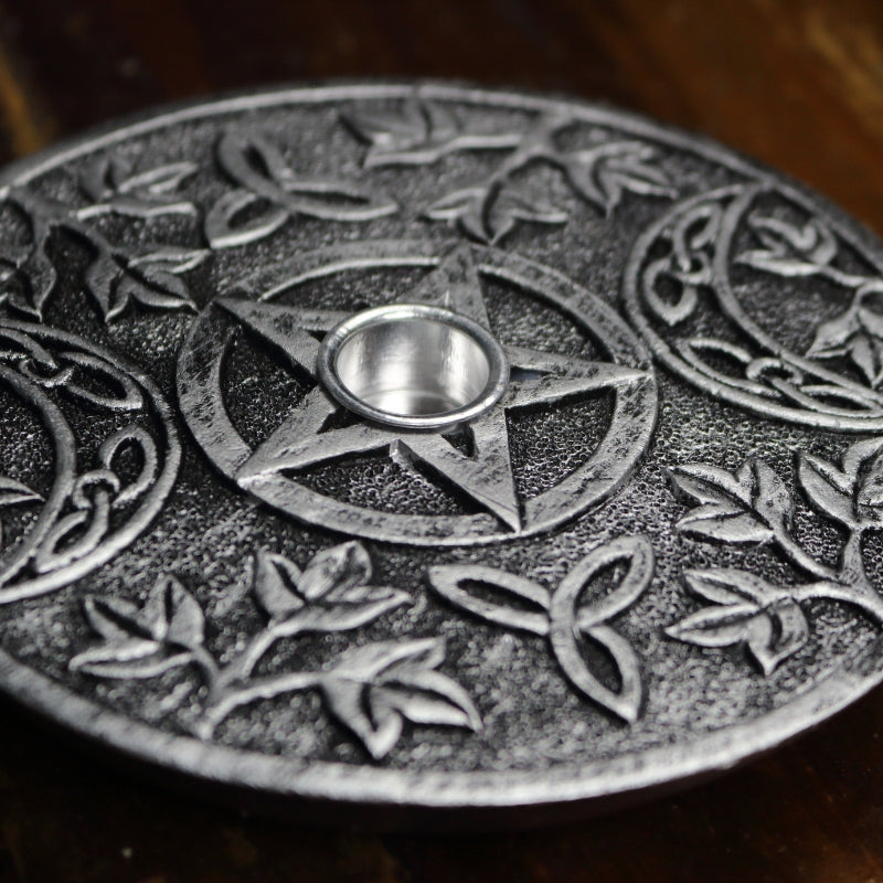 silver incense holder with an incense cone in the centre and incense stick behind with a pattern of a pentacle (5 pointed star) and 2 moons beside it. sitting on a wooden table