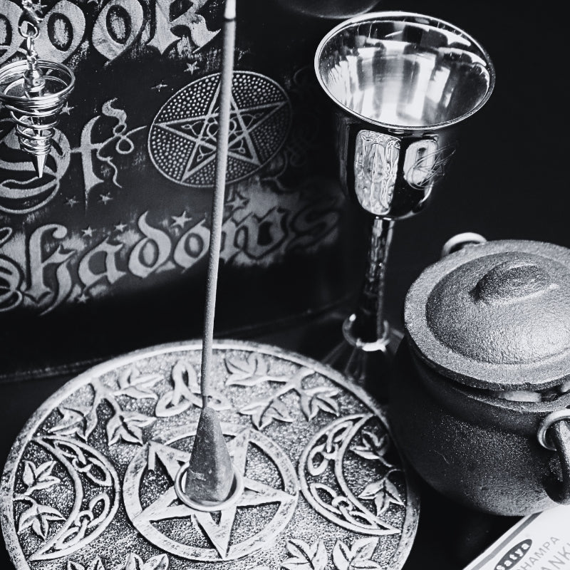 silver incense holder with an incense cone in the centre and incense stick behind with a pattern of a pentacle (5 pointed star) and 2 moons beside it. sitting on a black table with a brown box of frankincense incense, a silver chalice, black book of shadows and a cast iron cauldron with a silver handle.