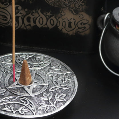 silver incense holder with an incense cone in the centre and incense stick behind with a pattern of a pentacle (5 pointed star) and 2 moons beside it. sitting on a black table with a brown box of frankincense incense, a silver chalice, black book of shadows and a cast iron cauldron with a silver handle.