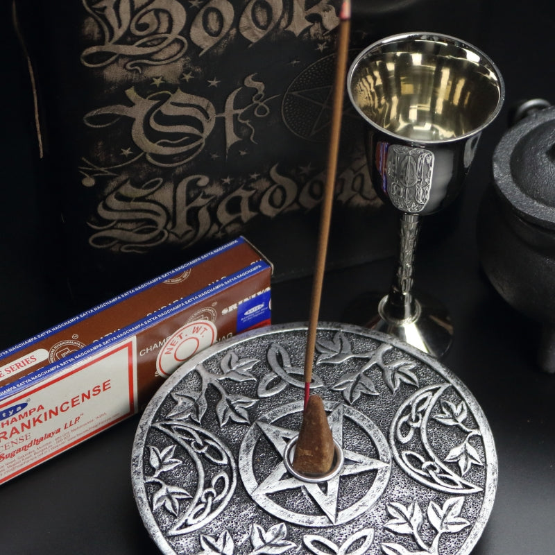 silver incense holder with an incense cone in the centre and incense stick behind with a pattern of a pentacle (5 pointed star) and 2 moons beside it. sitting on a black table with a brown box of frankincense incense, a silver chalice, black book of shadows and a cast iron cauldron with a silver handle.