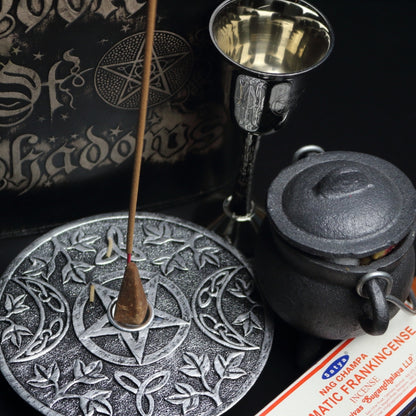 silver incense holder with an incense cone in the centre and incense stick behind with a pattern of a pentacle (5 pointed star) and 2 moons beside it. sitting on a black table with a brown box of frankincense incense, a silver chalice, black book of shadows and a cast iron cauldron with a silver handle.