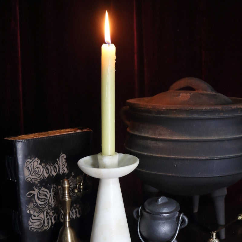 lit beeswax dinner candle in a white marble candle holder. In the background there is a large cauldron, a small cauldron , a black book of shadows , dark brass altar bell and an ornate brass candle snuffer