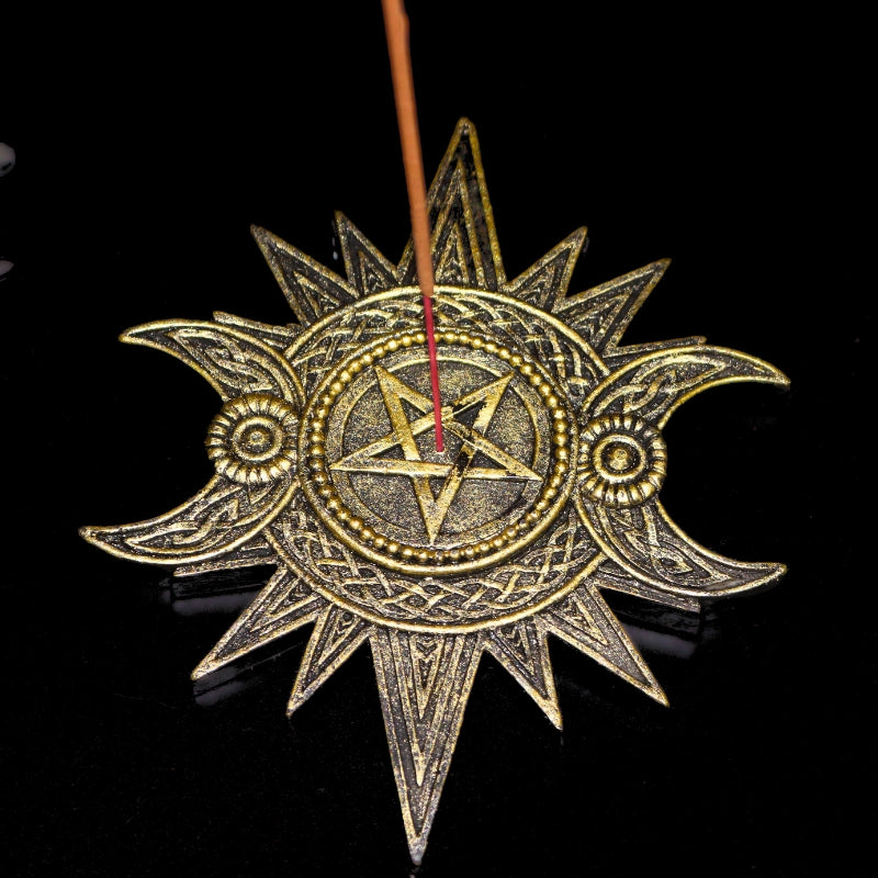 Gold coloured incense holder in the shape of a sun with a pentacle in the centre and two crescent moons at each side of the pentacle. An incense stick in the centre.