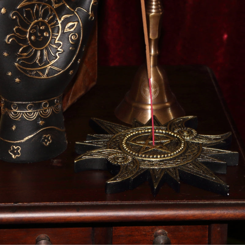 Gold coloured incense holder in the shape of a sun with a pentacle in the centre and two crescent moons at each side of the pentacle. An incense stick in the centre, sitting on top of a wooden apothecary cabinet, in front of a gold pentacle bell, next to a black and gold hand with sun and moon symbols on it.