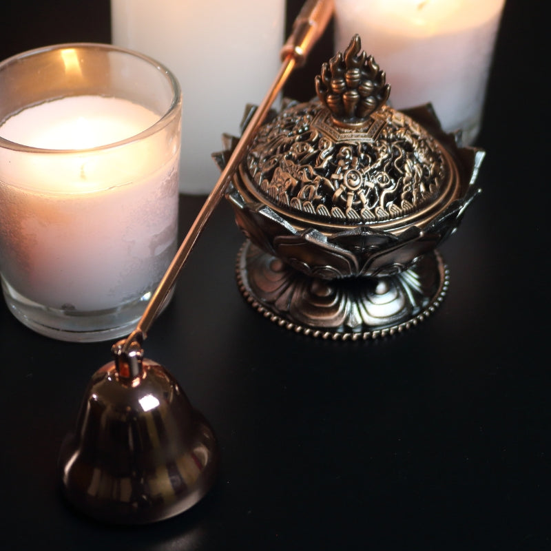 brass rose gold coloured candle snuffer  laid across an ornate brass incense holder, in front of 3 lit white candles on a black background