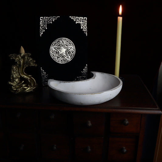 White washed carved crescent moon shaped bowl on an apothecary cabinet, next to a gold incense cone burner , black and silver velvet spell book and beeswax taper candle