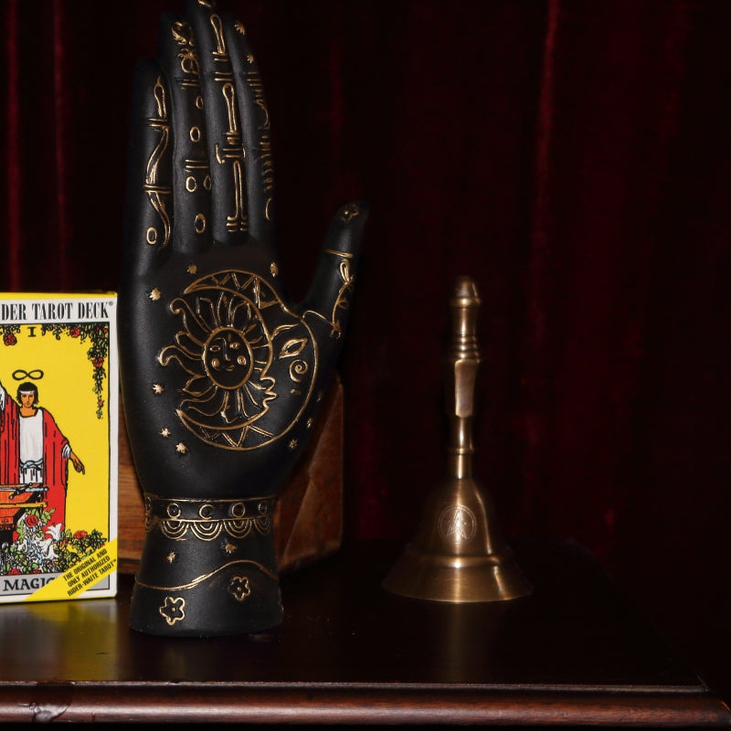 black and gold palmistry hand with sun and moon on the palm sitting on a wood apothecary cabinet next to a yellow pack of rider waite tarot cards and a bronze altar bell, all in front of a burgundy coloured curtain