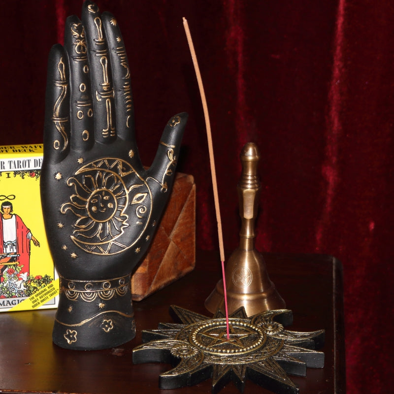 black and gold palmistry hand with sun and moon on the palm sitting on a wood apothecary cabinet next to a gold triple moon incense holder, a bronze altar bell, a yellow pack of rider waite tarot cards and a tarot box, all in front of a burgundy coloured curtain