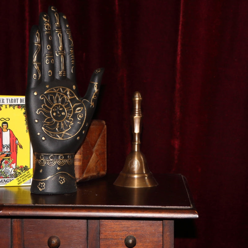 black and gold palmistry hand with sun and moon on the palm sitting on a wood apothecary cabinet next to a gold triple moon incense holder, a yellow pack of rider waite tarot cards and a tarot box, all in front of a burgundy coloured curtain