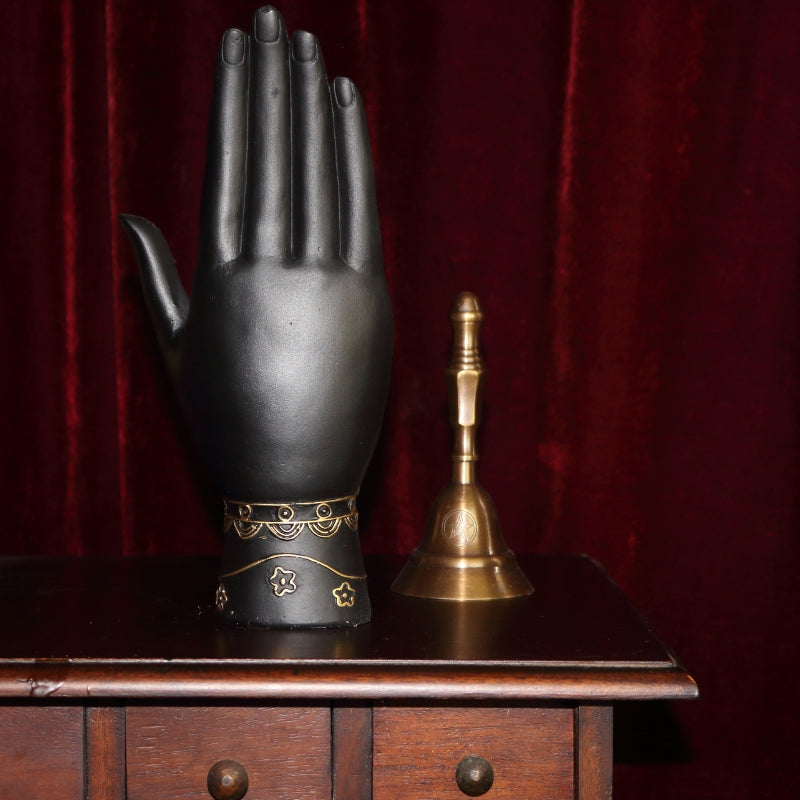 black and gold palmistry hand (back of hand) next to a bronze altar bell , all in front of a burgundy coloured curtain, sitting on a wooden apothecary cabinet
