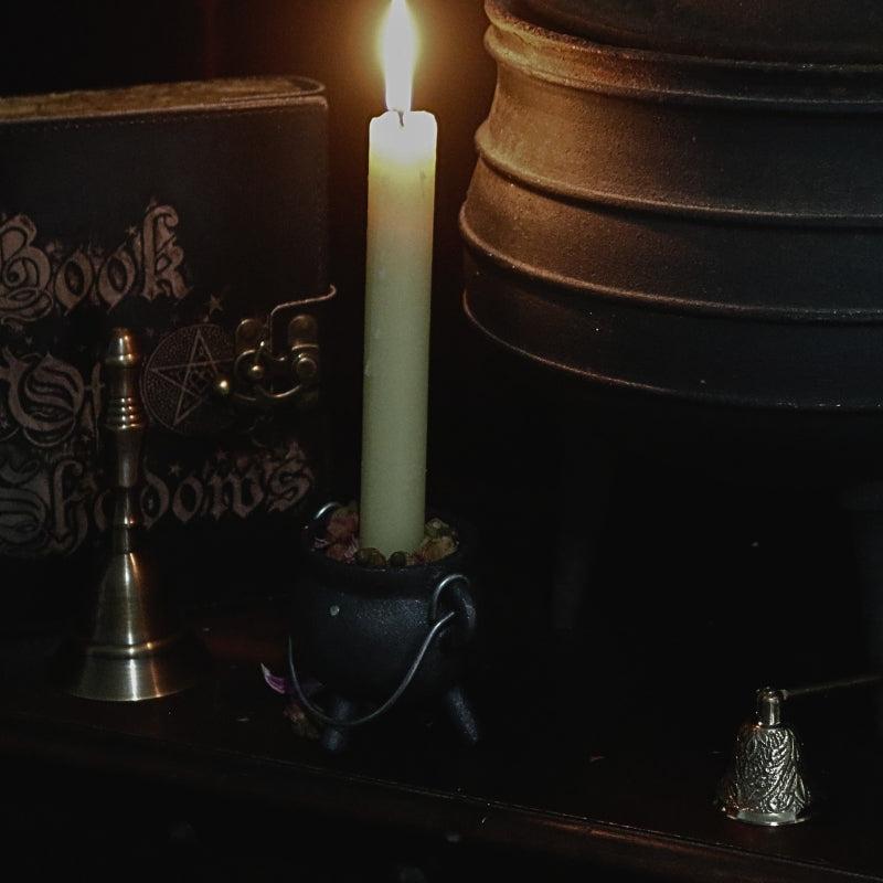lit beeswax dinner candle in a small cauldron. In the background there is a large cauldron, a black book of shadows , dark brass altar bell and an ornate brass candle snuffer