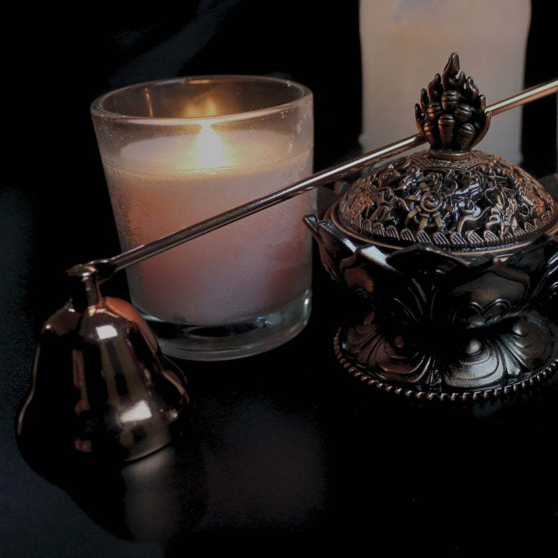 brass rose gold coloured candle snuffer  laid across an ornate brass incense holder, in front of  2 lit white candles on a black background