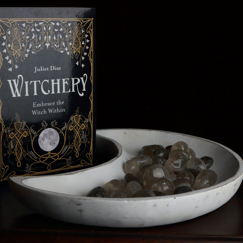 whitewash crescent moon shaped bowl filled with smoky quartz crystals in front of a juliet Diaz book called "Witchery"