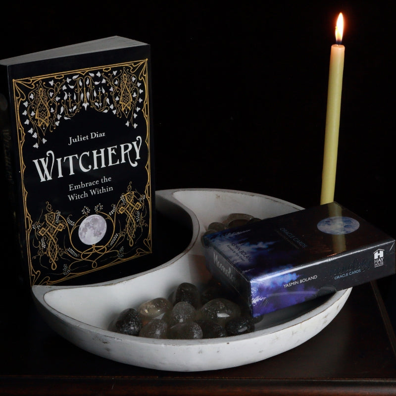 whitewash crescent moon shaped bowl filled with smoky quartz crystals and holding a moonology oracle deck,  in front of a juliet Diaz book called "Witchery" and a beeswax prayer candle