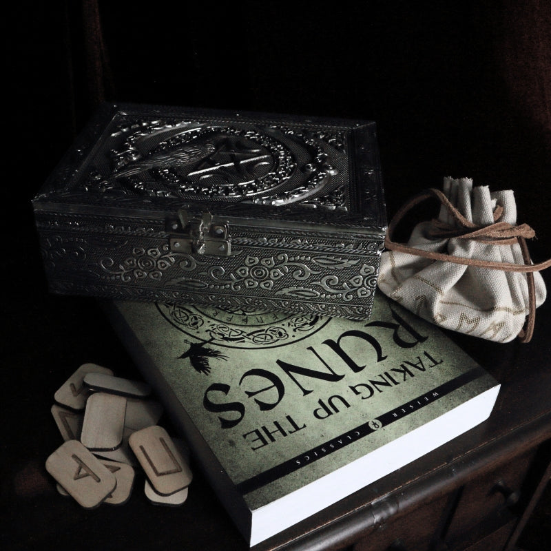 embossed metal tarot box with a raven on top, sitting on a book about runes, with wooden runes surrounding 