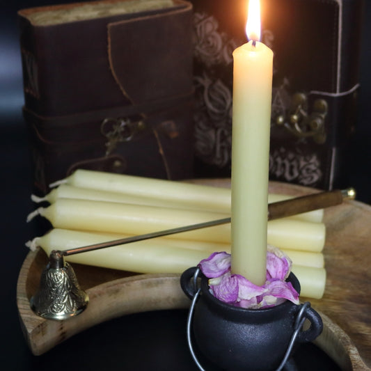 lit beeswax candle sitting in a cauldron, in front of a wooden crescent moon shaped bowl with a brass embellished candle snuffer and old leather journals