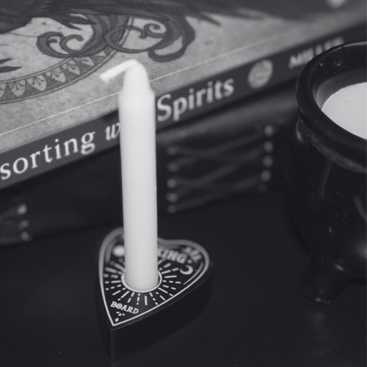 Black spell candle holder shaped as a Ouija board planchette, holding  a white spell candle. In front of a book "consorting with spirits" , leather journal and black ceramic cauldron candle