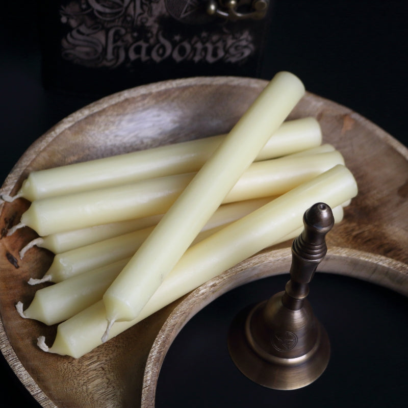 beeswax taper candles sitting in a wooden carved crescent moon shaped bowl next to an altar bell, in front of a leather book of shadows