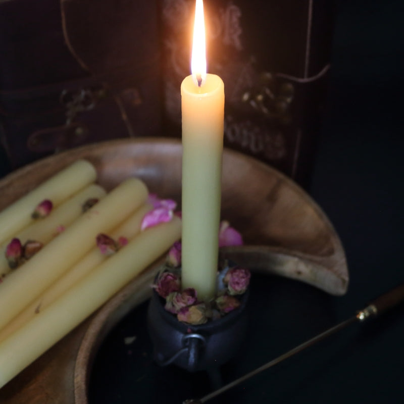 lit beeswax candle sitting in a cauldron, in front of a wooden crescent moon shaped bowl with a brass embellished candle snuffer and old leather journals
