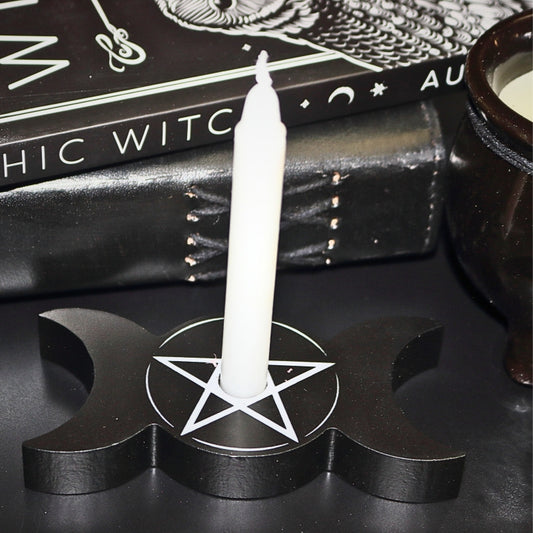 Black triple moon shaped candle holder with pentacle design in white. Holding a white spell candle, in front of the psychic witch book, leather journal and black cauldron candle