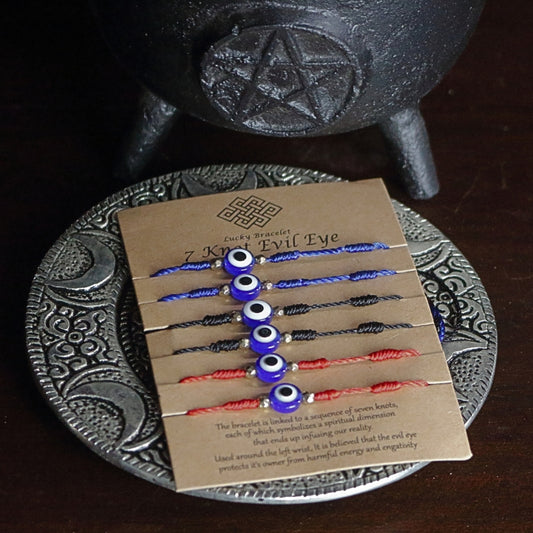 brown cardboard card with 6 corded bracelets, 2 red, 2 blue and 2 black, each with 7 knots and one nazar evil eye bead in the centre. Sitting on a silver embossed plate in front of a black cast iron cauldron with a pentacle on the front.