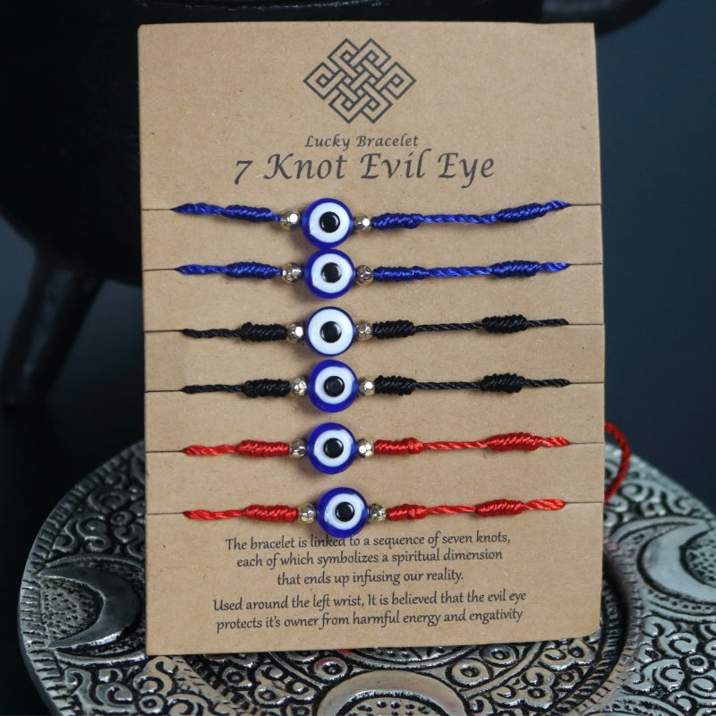 brown cardboard card with 6 corded bracelets, 2 red, 2 blue and 2 black, each with 7 knots and one nazar evil eye bead in the centre. Sitting on a silver embossed plate in front of a black cast iron cauldron with a pentacle on the front.