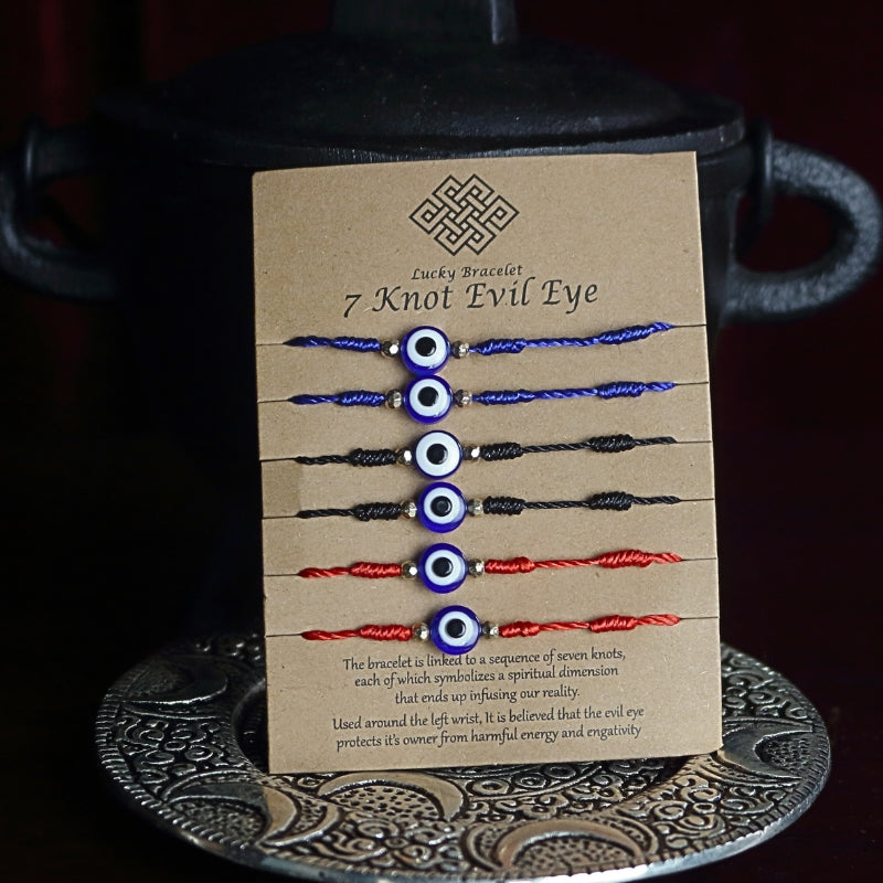 brown cardboard card with 6 corded bracelets, 2 red, 2 blue and 2 black, each with 7 knots and one nazar evil eye bead in the centre. Sitting on a silver embossed plate in front of a black cast iron cauldron with a pentacle on the front.