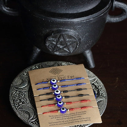 brown cardboard card with 6 corded bracelets, 2 red, 2 blue and 2 black, each with 7 knots and one nazar evil eye bead in the centre. Sitting on a silver embossed plate in front of a black cast iron cauldron with a pentacle on the front.