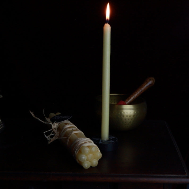 bundle of beeswax prayer candles next to a lit beeswax candle and a brass singing bowl, on a wooden apothecary cabinet