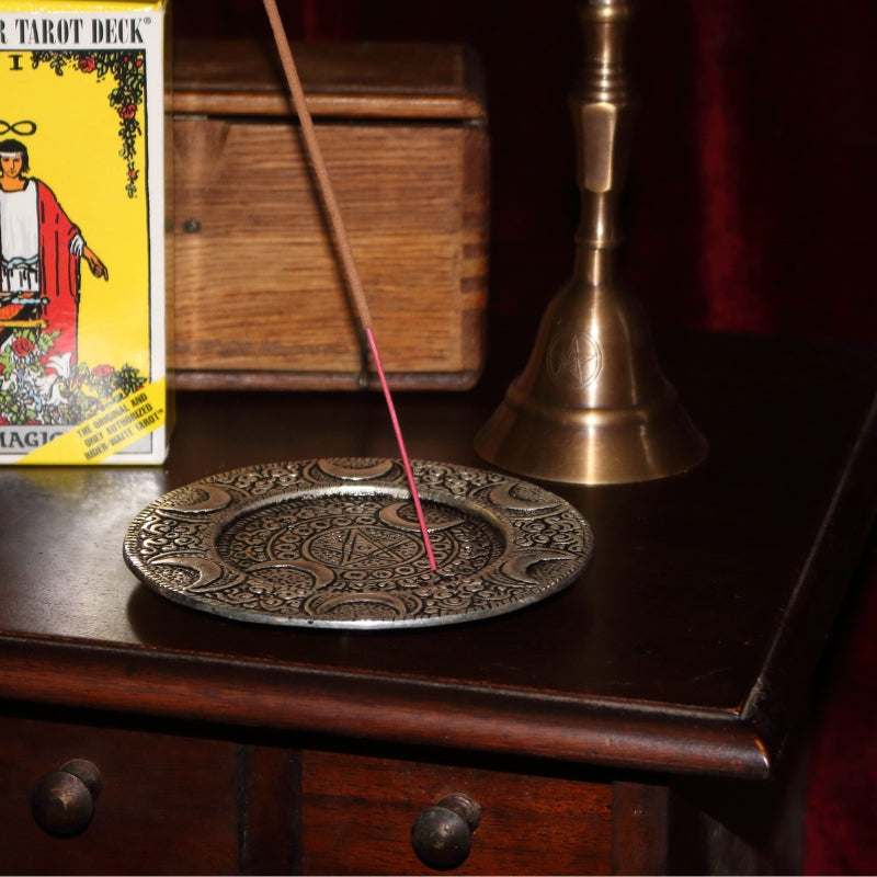 silver triple moon incense holder with incense stick, sitting on a wooden apothecary cabinet in front of a brass altar bell, rider waite tarot deck and wooden box