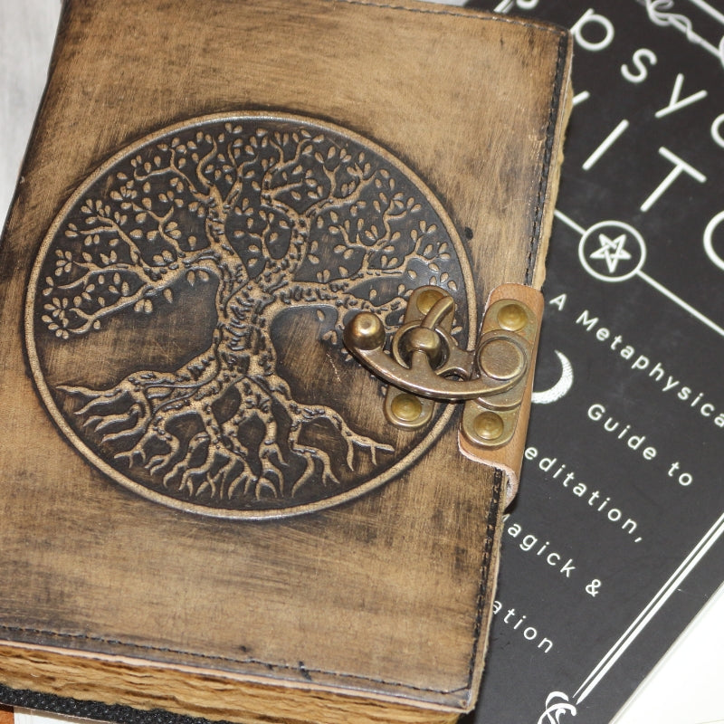 Antique style brown leather journal with the tree of life on the cover and a brass closure. Inside contains plain "antiqued" pages . Sitting on a copy of mat auryn's "Psychic Witch" book