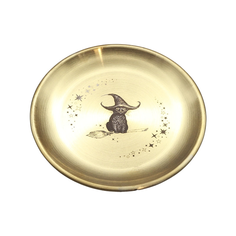 A round, brushed gold metal tray or shallow dish featuring a central illustration of a black owl wearing a witch's hat on a broomstick, surrounded by small stars.