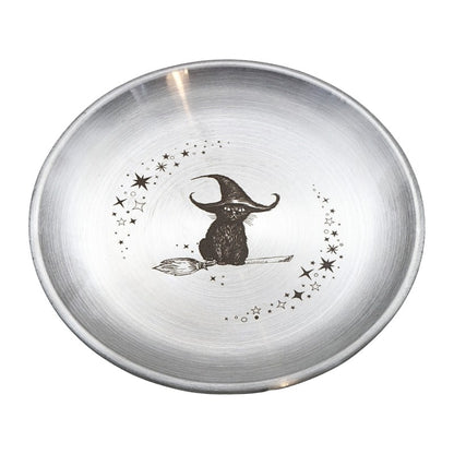 A round brushed silver metal bowl featuring a black illustration of a witch sitting on a broomstick, surrounded by stars and dots, on a white background.