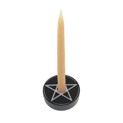 A single cream-colored bees wax taper candle standing upright in a round black holder engraved with a white pentagram, set against a plain white background.