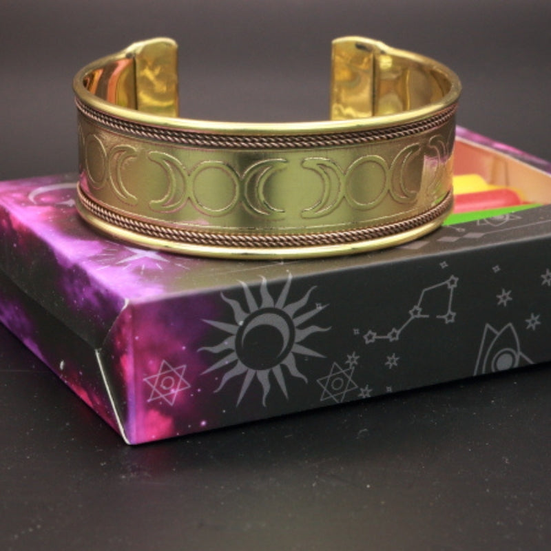 gold triple moon design cuff bracelet sitting atop a box of mixed coloured spell candles