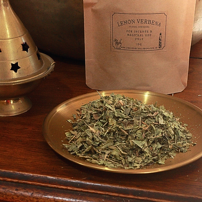 A kraft paper pouch labeled 'LEMON VERBENA' stands behind a brass plate filled with dried green herb leaves on a wooden surface.