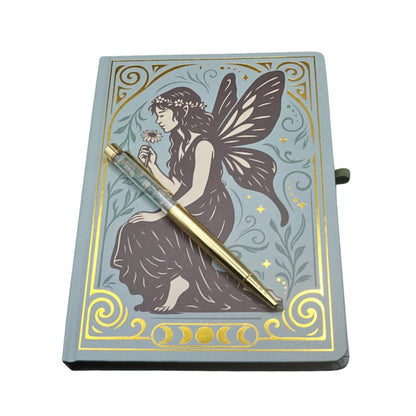 blue and gold notebook with a grey vintage style fairy on the cover, and a gold pen on top, on a white background