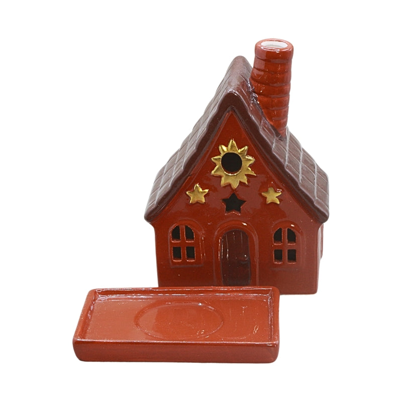 A small decorative house-shaped ceramic container or figurine with a red body, brown roof, red-brown door, and gold sun and star emblems next to a rectangular base a plain white background.