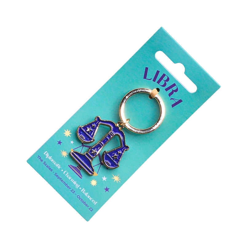 A Libra-themed keychain charm featuring a purple and blue enamel scales of justice design with a gold-tone keyring, attached to a turquoise card printed with the word 'LIBRA'.