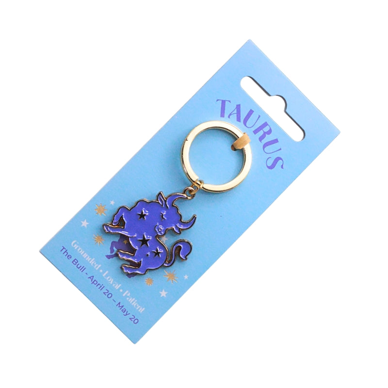 A Taurus zodiac sign keychain featuring a purple bull with star details and a gold-tone key ring, attached to a light blue cardboard display card labeled 'TAURUS'.