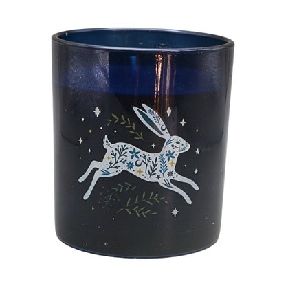 A dark navy blue glass candle jar featuring a white line-art illustration of a hopping rabbit decorated with blue and green floral patterns, surrounded by small stars and leaves.